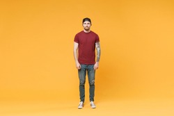 Full body length handsome young tattooed man in basic print t-shirt black cap standing looking camera isolated on yellow wall background studio portrait. People lifestyle concept. Mock up copy space