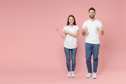 Full body length young cheerful couple two friends man woman in white basic blank print design t-shirts jeans standing pointing aside workspace isolated on pastel pink color background studio portrait