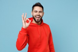 Young caucasian smiling happy bearded attractive handsome positive man 20s wearing casual red orange hoodie show ok okay gesture isolated on blue background studio portrait People lifestyle concept
