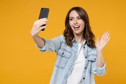 Young friendly fun woman 20s wearing stylish denim shirt white t-shirt doing selfie shot on mobile cell phone talking by video call waving hand greeting isolated on yellow background studio portrait.