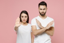 Young couple two friends bearded man woman in white basic blank print design t-shirts say no holding palm folded crossed hands in stop gesture isolated on pastel pink color background studio portrait