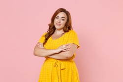 Smiling beautiful young redhead plus size body positive female woman girl 20s in yellow dress posing holding hands crossed looking camera isolated on pastel pink color wall background studio portrait
