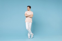 Full length of young caucasian attractive fashionable student man 20s wearing beige t-shirt white pants holding hands crossed folded looking aside isolated on blue color background studio portrait