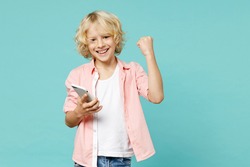 Happy little kid boy 10s wearing pink shirt using mobile cell phone typing sms message doing winner gesture isolated on blue turquoise background children studio portrait. Childhood lifestyle concept