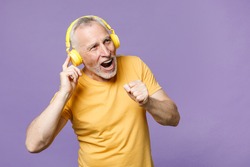 Cheerful elderly gray-haired mustache bearded man in casual yellow t-shirt posing isolated on violet wall background studio portrait. People lifestyle concept. Listen music with headphones, sing song