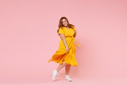 Full length portrait of smiling beautiful charming young redhead plus size body positive female woman girl 20s in yellow dress posing looking camera isolated on pastel pink color background studio
