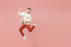 Full length young smiling happy overjoyed joyful fun trendy caucasian excited man 20s wearing jacket white t-shirt jump high playing guitar isolated on pastel pink color background studio portrait.