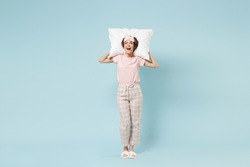 Full length young excited happy woman 20s in pajamas jam sleep eye mask rest relax at home holding pillow look aside isolated on pastel blue color background studio. Good mood night bedtime concept.