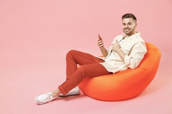 Full length young happy fun man in jacket white t-shirt sitting in bean bag chair using mobile cell phone chat online in social network show thumb up gesture isolated on pastel pink background studio.