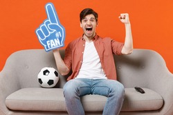 Young excited man football fan in shirt foam glove finger support favorite team with soccer ball sit home sofa watch tv live stream do winner gesture isolated on orange background People sport concept