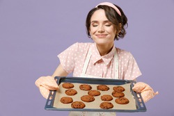 Young housewife housekeeper chef cook baker woman wearing pink apron showing chocolate cookies biscuits on baking sheet sniff chocolate smell isolated on pastel violet background. Cooking food concept