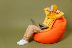 Full length young arabian asian muslim woman in abaya hijab yellow clothes hold laptop pc computer sit bean bag chair rest relax isolated on olive green background. People uae islam religious concept