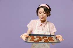 Young excited happy housewife housekeeper chef cook baker woman wearing pink apron showing chocolate cookies biscuits on baking sheet isolated on pastel violet background studio. Cooking food concept.