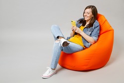 Full length young fun caucasian woman wear denim jacket yellow t-shirt use mobile cell phone browsing sitting in bean bag chair isolated on grey background studio portrait. People lifestyle concept.
