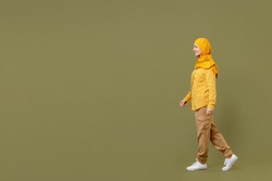 Full length side view young arabian asian muslim woman 20s in abaya hijab yellow clothes walking going strolling isolated on olive green background. People uae middle eastern islam religious concept.