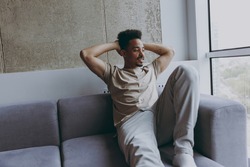 Dreamful young african american man in beige t-shirt sweatpants sit on comfortbale grey sofa indoors apartment look aside at window hold hands behind neck procrastinate, rest on weekends stay at home.