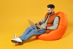 Full length young smiling freelancer fun caucasian man 20s years old wear orange vest mint sweatshirt sitting in beanbag bag chair hold laptop pc computer isolated on yellow background studio portrait