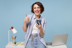 Young happy fun secretary employee business woman in casual shirt work stand at white office desk with pc laptop hold mobile cell phone show thumb up gesture isolated on pastel blue background studio.