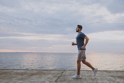 Side view young strong sporty athletic toned fit sportsman man 20s in sports clothes warm up training run jogging at sunrise sun over sea beach outdoor on pier seaside in summer day cloudy morning.