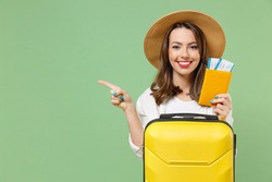 Close up tourist woman in casual clothes hat hold passport tickets yellow suitcase point finger aside workspace isolated on green background Passenger travel abroad weekend getaway Air flight concept.