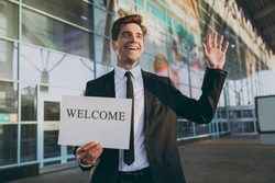 Bottom view young cheerful traveler businessman man in black suit stand outside at international airport terminal hold card sign with welcome title text waving hand Air flight business trip concept.