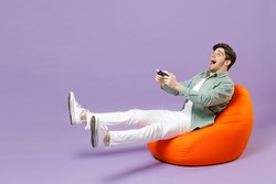 Full length man in casual mint shirt white t-shirt sitting in orange bean bag chair hold takeaway bucket eat popcorn watch movie film play pc game with joystick console isolated on purple background.