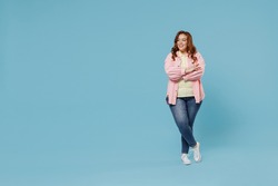 Full length young fun smiling redhead chubby overweight woman 30s wearing in pink shirt jeans casual clothes hold hands crossed folded look aside isolated on pastel blue background studio portrait.