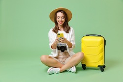 Full length traveler fun tourist woman in casual clothes hat hold suitcase using mobile cell phone sit isolated on green background Passenger travel abroad weekends getaway Air flight journey concept