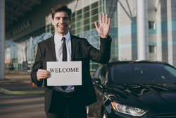 Bottom view young friendly traveler businessman man wear black suit stand outside at international airport terminal hold card sign with welcome title text waving hand Air flight business trip concept.