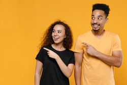 Young couple two friends together family smiling happy african man woman 20s wearing black t-shirt point index finger aside on workspace area copy space isolated on yellow background studio portrait