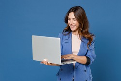 Smiling cheerful funny beautiful attractive young brunette woman 20s wearing basic casual jacket standing working on laptop pc computer isolated on bright blue colour background studio portrait