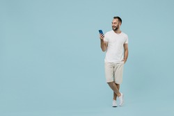 Full length young smiling friendly happy man 20s wearing casual white t-shirt looking camera using mobile cell phone chat onine isolated on plain pastel light blue color background studio portrait.