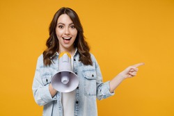 Young expressive fun woman 20s wear denim shirt white t-shirt screaming hot news shouting aside in megaphone point index finger aside on workspace area isolated on yellow background studio portrait.