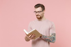 Handsome young bearded tattooed man guy in pastel casual t-shirt, eyeglasses isolated on pink background studio portrait. People sincere emotions lifestyle concept. Mock up copy space. Reading book