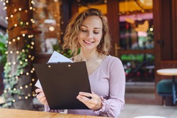 Young happy smiling waiter businesswoman woman 20s wear casual clothes look aside standing at coffee shop cafe hold black clipboard with papers reading document menu working in restaurant indoors.