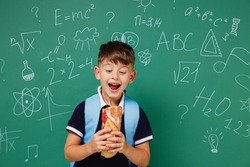 Young amazed male kid school boy 5-6 years old in t-shirt backpack biting sandwich eating lunch isolated on green wall chalk blackboard background. Childhood children kids education lifestyle concept.