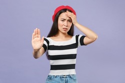Worried dissatisfied young asian woman in striped t-shirt red beret standing showing stop gesture with palm put hand on head looking camera isolated on pastel violet colour background studio portrait
