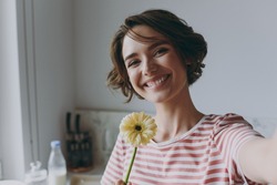 Close up young housewife woman 20s in casual clothes striped t-shirt holding flower gerbera doing selfie shot on mobile phone cooking food in light kitchen at home alone Healthy diet lifestyle concept