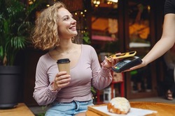 Young smiling woman 20s in casual clothes at cafe buy breakfast sit at table hold wireless bank payment terminal mobile phone to process acquire payments relax in restaurant during free time indoors.
