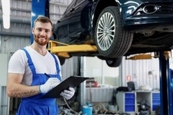 Young happy technician mechanic man wears denim overalls use hold clipboard papers document writing estimate stand near car lift check technical condition work in vehicle repair shop workshop indoors