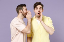 Two shocked young men friends together in casual t-shirt tattoo translate fun whisper gossip and tell secret behind his hand sharing news scream isolated on purple background. People lifestyle concept