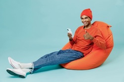 Full body young smiling fun happy african american man wearing orange shirt hat sit in bag chair use hold mobile cell phone show thumb up gesture isolated on plain pastel light blue background studio