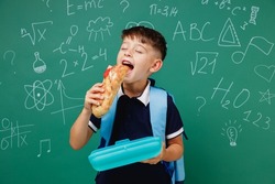 Young male kid school boy 5-6 years old in t-shirt backpack biting sandwich eat lunch at break isolated on green wall chalk blackboard background. Childhood children kids education lifestyle concept.