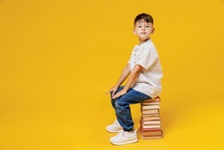 Side view young smart wunderkind school boy 5-6 years old in t-shirt casual clothes sit on pile of books isolated on plain yellow background studio Childhood children kids education lifestyle concept