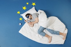 Full length top view young man in pajamas jam sleep mask relax at home lies wrap covered under blanket hold teddy bear plush toy isolated on dark blue sky background. Good mood night bedtime concept