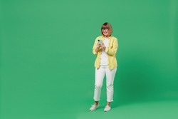 Full body elderly smiling cool happy woman 50s in glasses yellow shirt using hold in hand mobile cell phone browse internet isolated on plain green background studio portrait People lifestyle concept