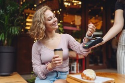 Young woman in casual clothes at cafe buy breakfast sit at table hold wireless bank payment terminal smart watch to process acquire payments drink coffee relax in restaurant during free time indoors.