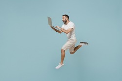 Full length side view young overjoyed man 20s in casual white t-shirt jump high run fast hurrying up hold closed laptop pc computer isolated on plain pastel light blue color background studio portrait