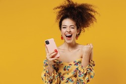 Young happy beautiful overjoyed woman 20s with culry hair in casual clothes hold use mobile cell phone do winner gesture isolated on plain yellow background studio portrait People lifestyle concept