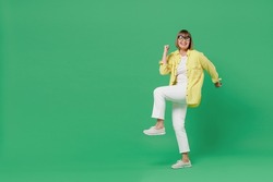 Full body elderly smiling happy overjoyed cool fun woman 50s in glasses yellow shirt do winner gesture clench fist celebrate isolated on plain green background studio portrait People lifestyle concept